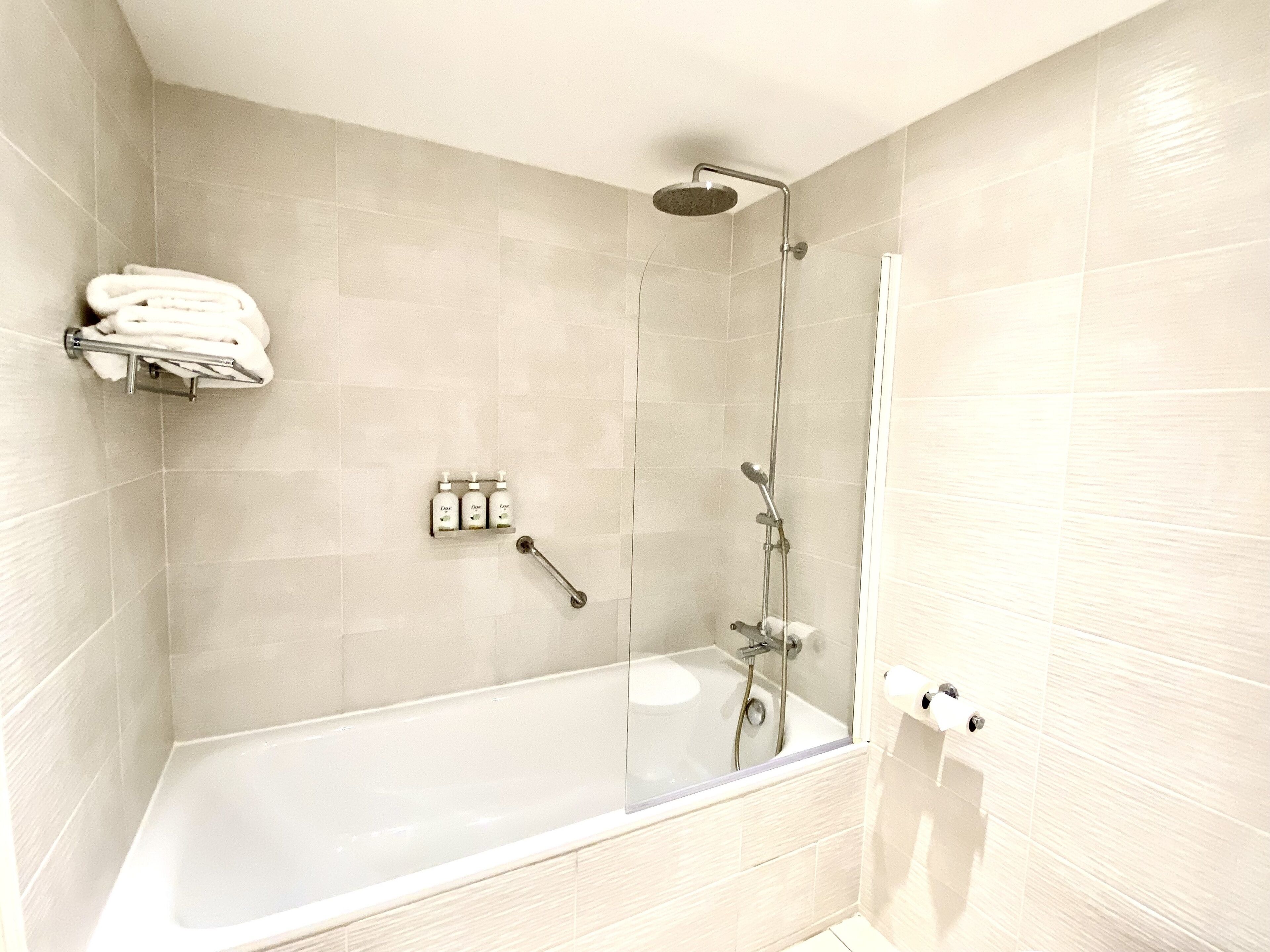 A modern bathroom with a glass-enclosed bathtub, a handheld showerhead, and a towel rack with folded towels.