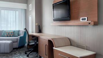 Une chambre d’hôtel avec un bureau en bois, un téléviseur fixé au mur, un canapé agrémenté d’un coussin à motifs et un sol recouvert de moquette.