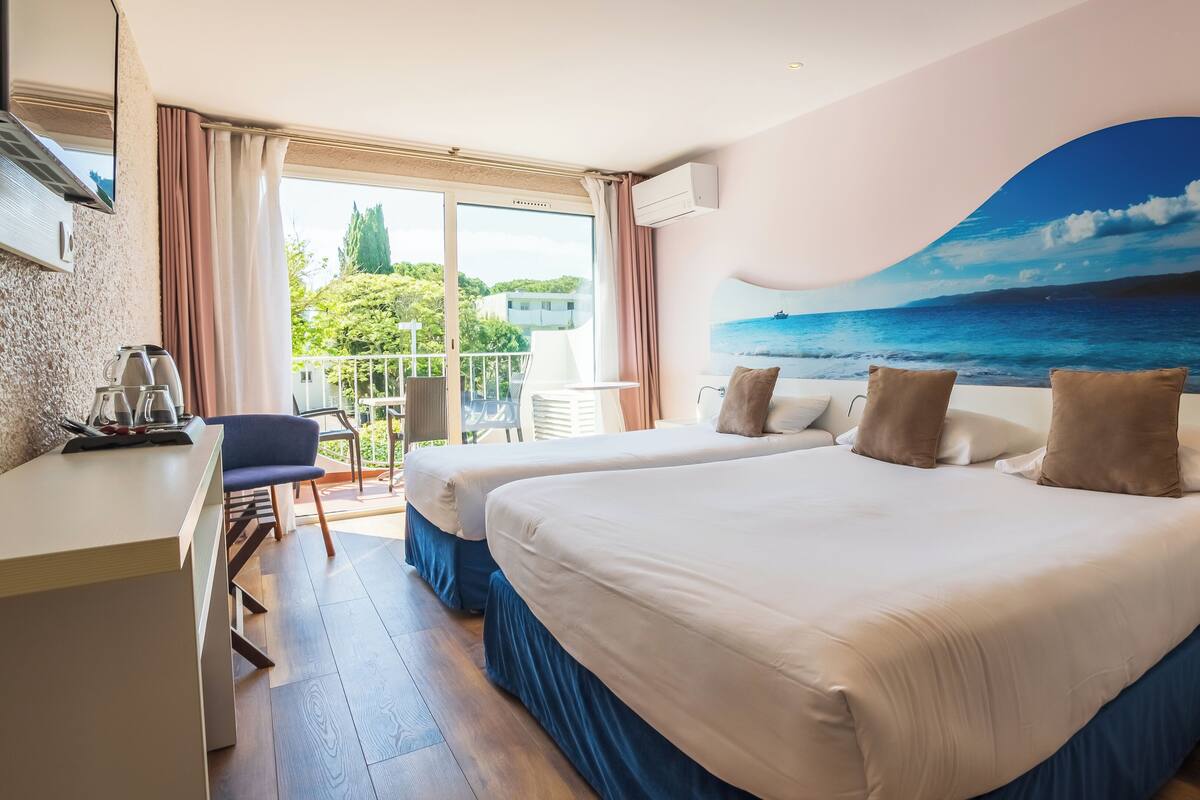 A hotel room with two beds, a desk with a chair, a balcony with a view of the sea, and an air conditioning unit.