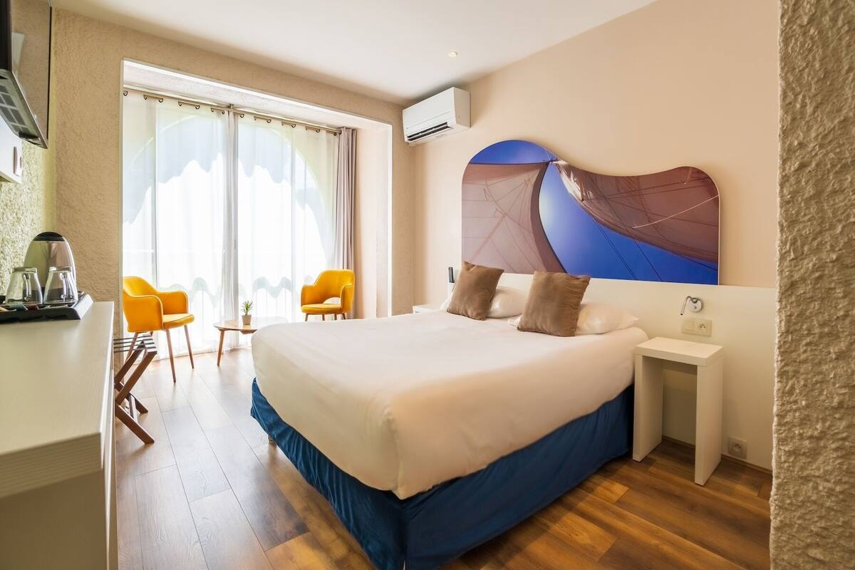 A hotel room with a large bed, a desk with a coffee maker, two yellow chairs, and a large window with curtains.
