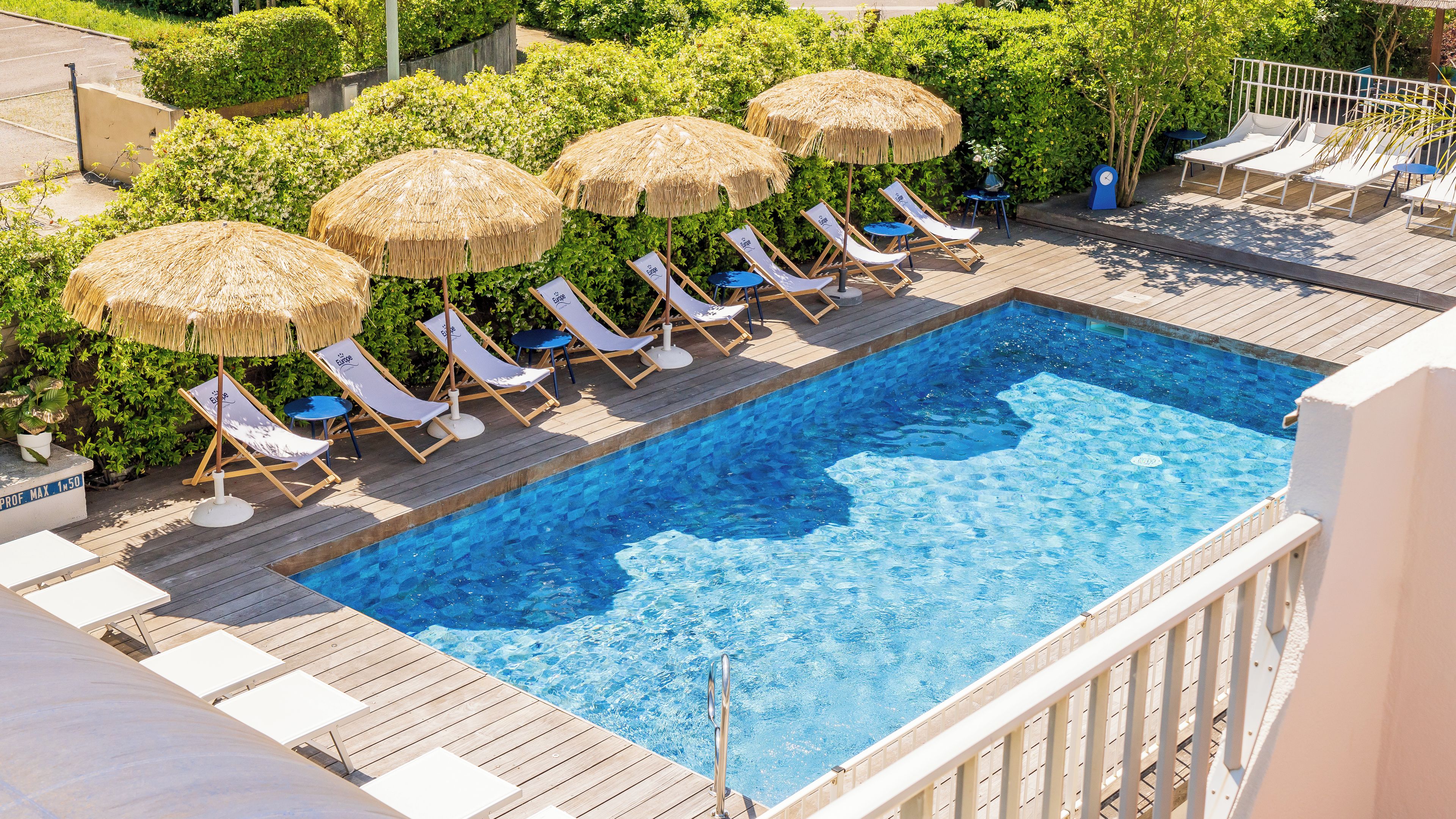 Una piscina con sillones de descanso y sombrillas de paja, rodeada de vegetación.