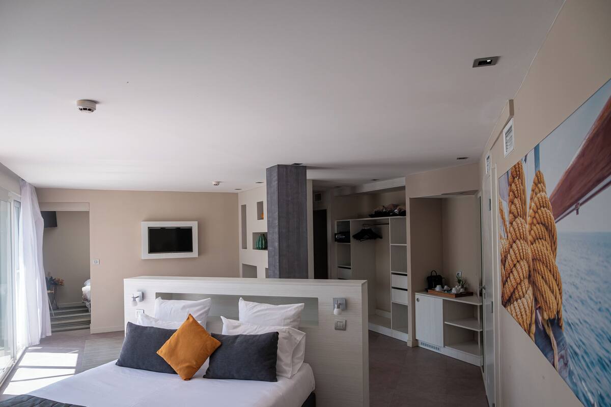 A modern hotel room with a large bed, a TV mounted on the wall, a closet, and a view of the sea through the window.