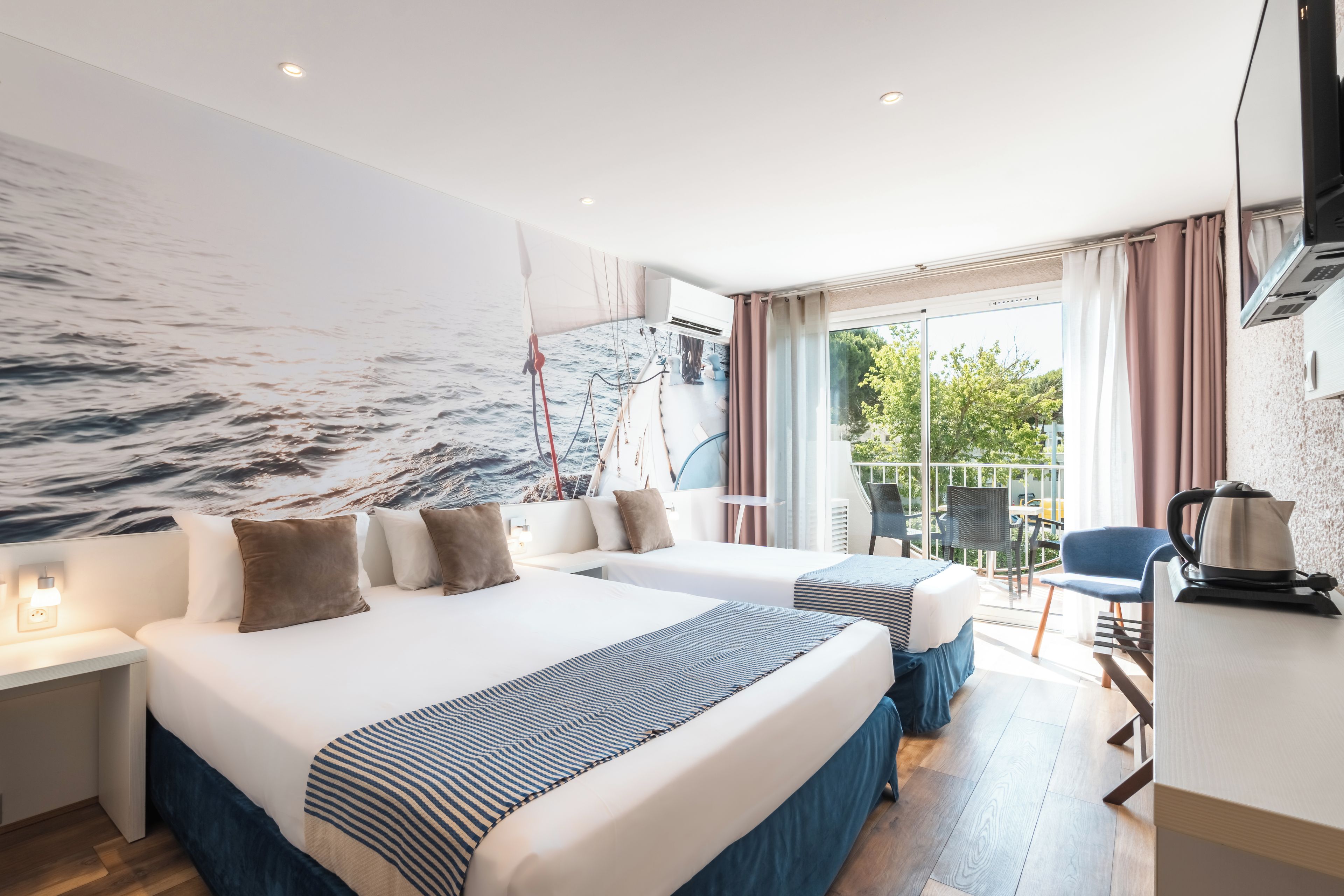 Habitación de hotel con dos camas, un ventanal con cortinas, vista al mar y un balcón con mobiliario exterior.