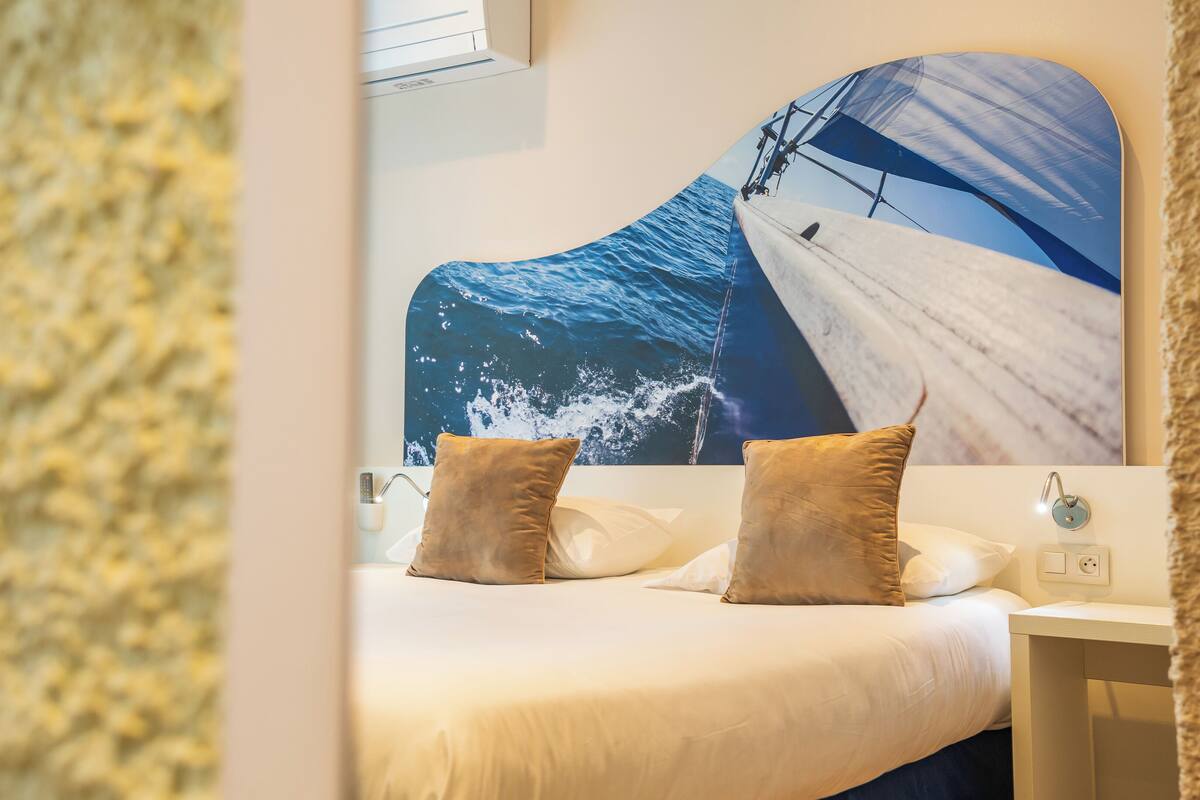A hotel room with a large ocean-themed mural on the wall, a bed with white bedding and two pillows, a small bedside table with a lamp, and a wall-mounted air conditioning unit.