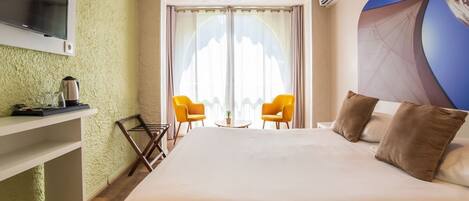 Una habitación de hotel con una cama, dos sillas amarillas, un televisor, una tetera y una ventana con cortinas.