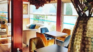 A modern lounge area with a wooden floor, blue and brown upholstered chairs, a central table, and a large vase with red flowers. - Hôtel Europe (La Grande-Motte)