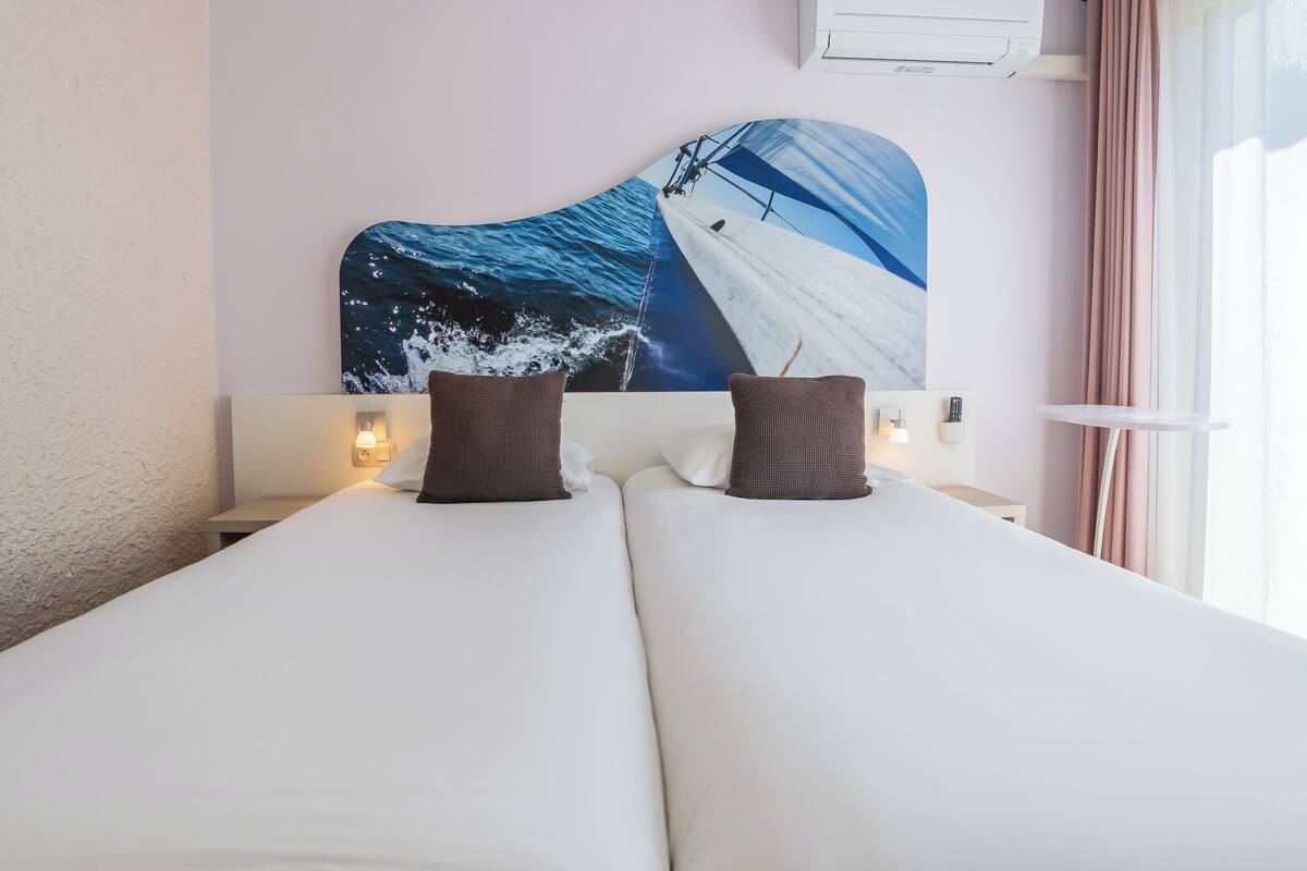 A double bed with white linens, two brown pillows, a nightstand with a lamp, and a wall-mounted air conditioner.