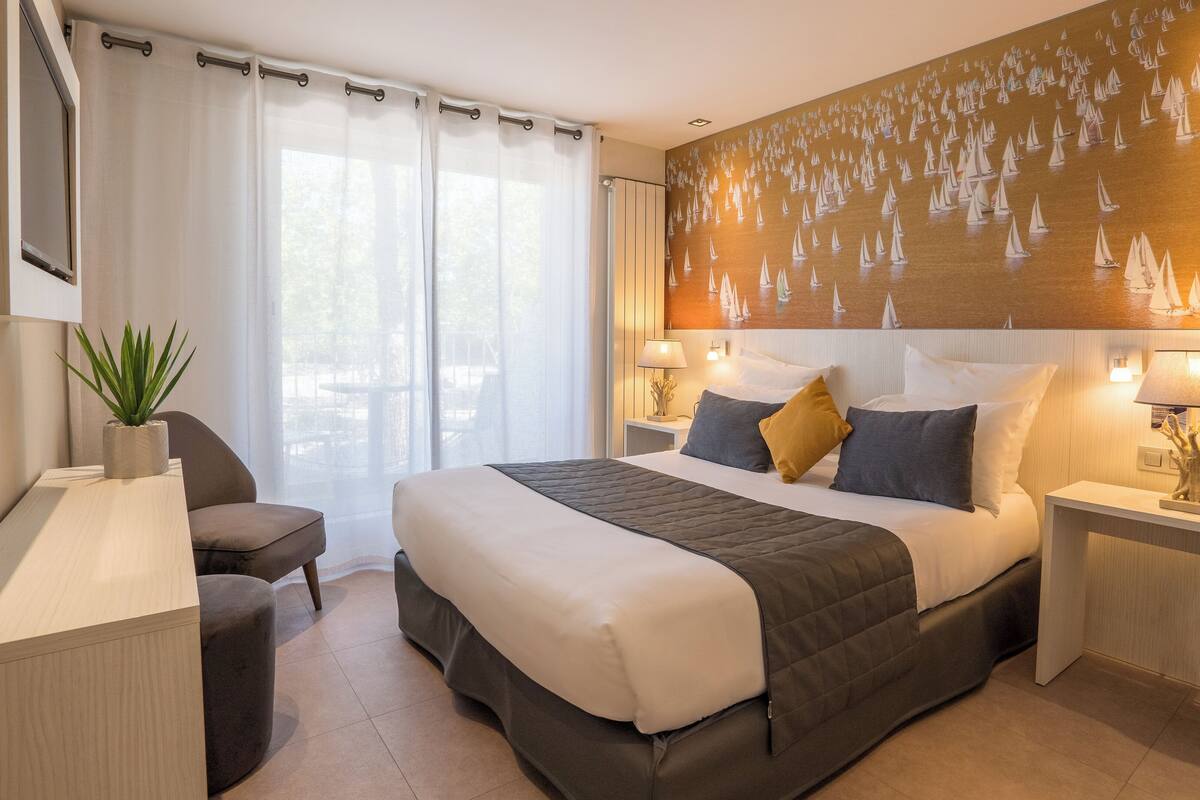 A hotel room with a bed, bedside tables, a desk, a chair, and a large sailboat mural on the wall.