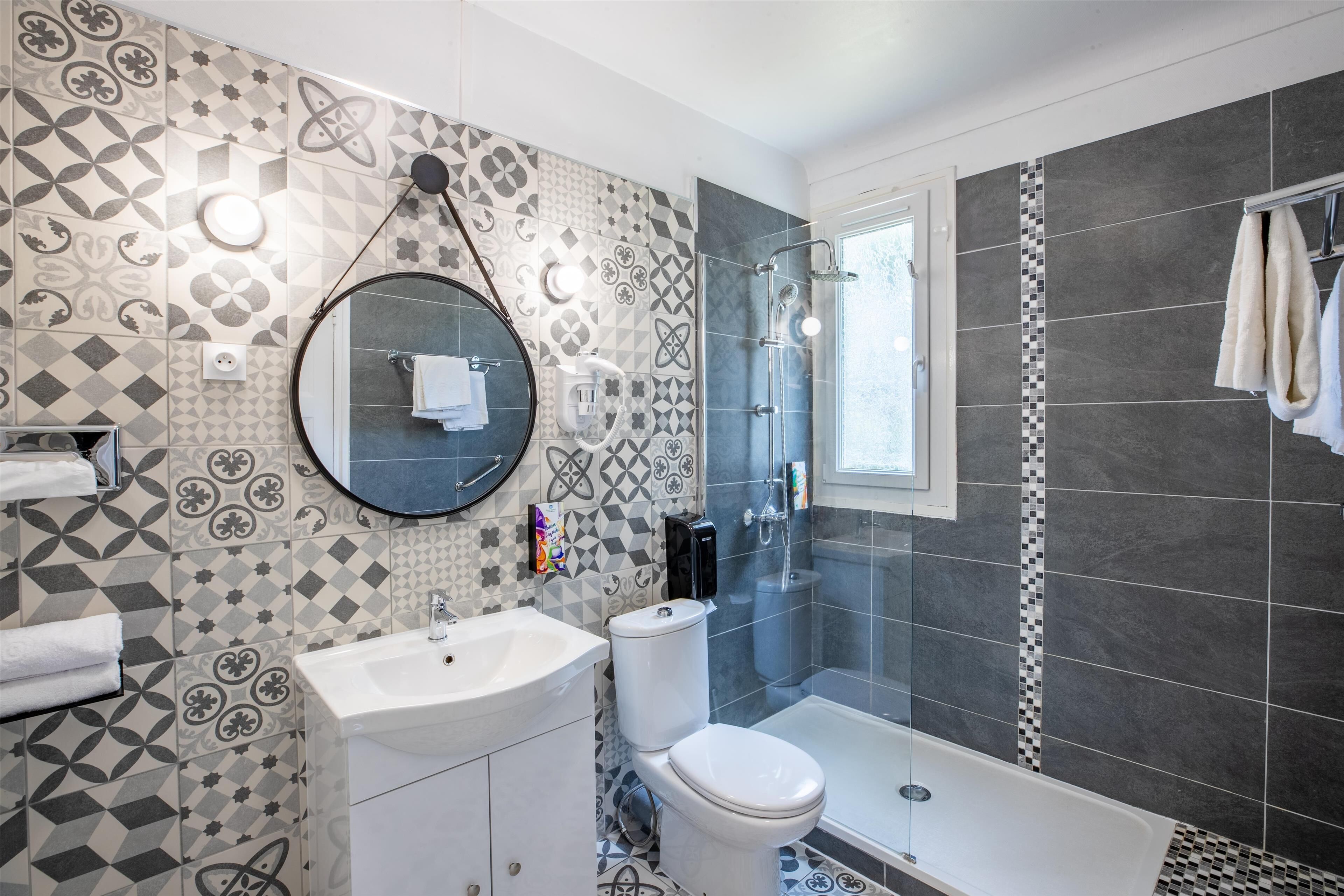 A modern bathroom with a geometric tile backsplash, a round mirror, a toilet, a sink, and a shower area.