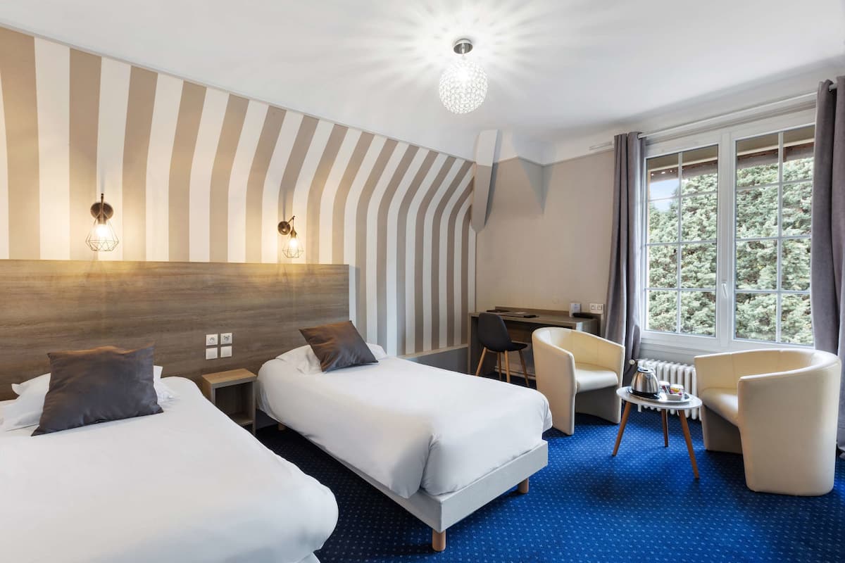 A hotel room with two beds, a desk, a chair, a small table with a kettle, and a large window with curtains.