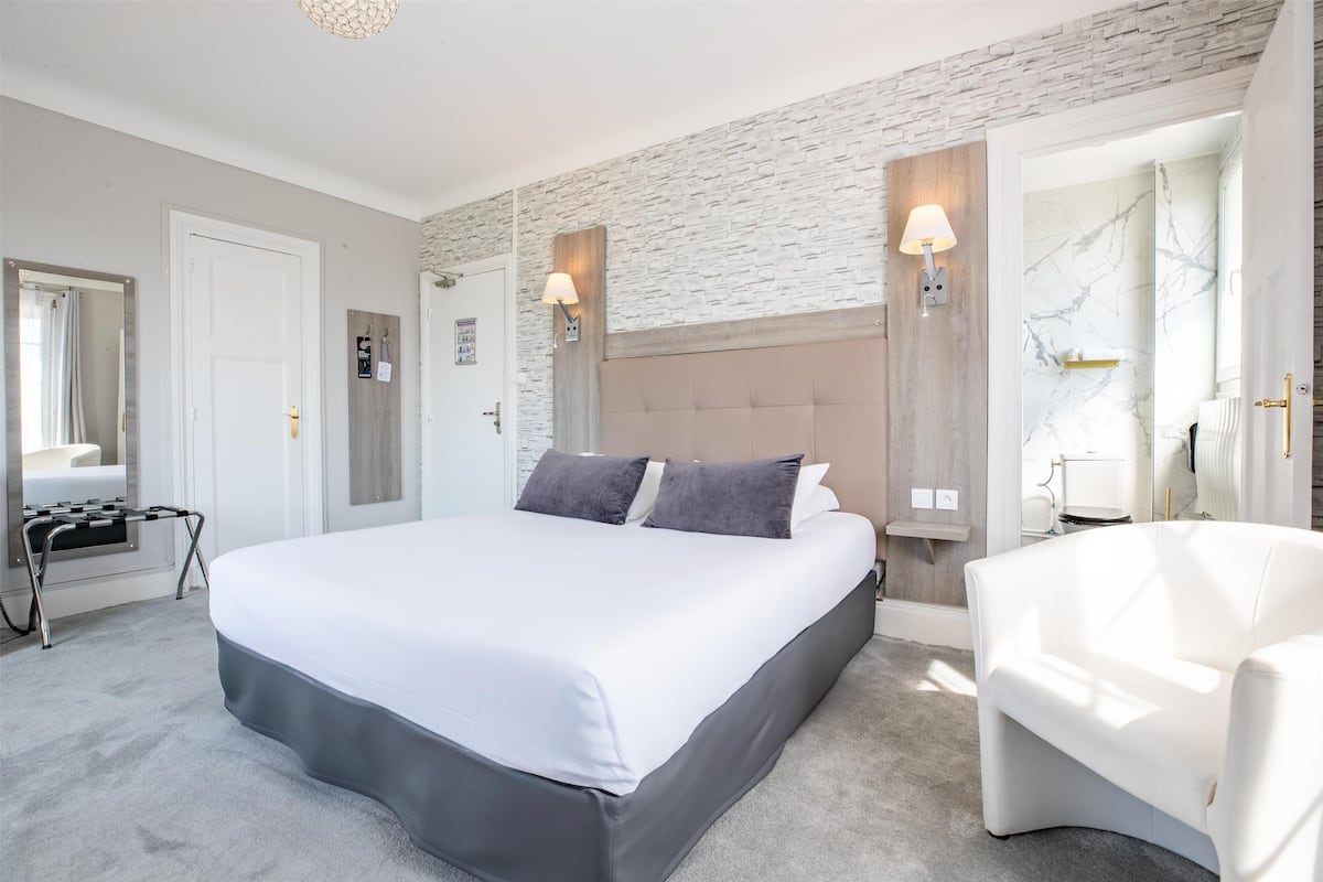 A modern hotel room with a large bed, a white armchair, and a brick accent wall.