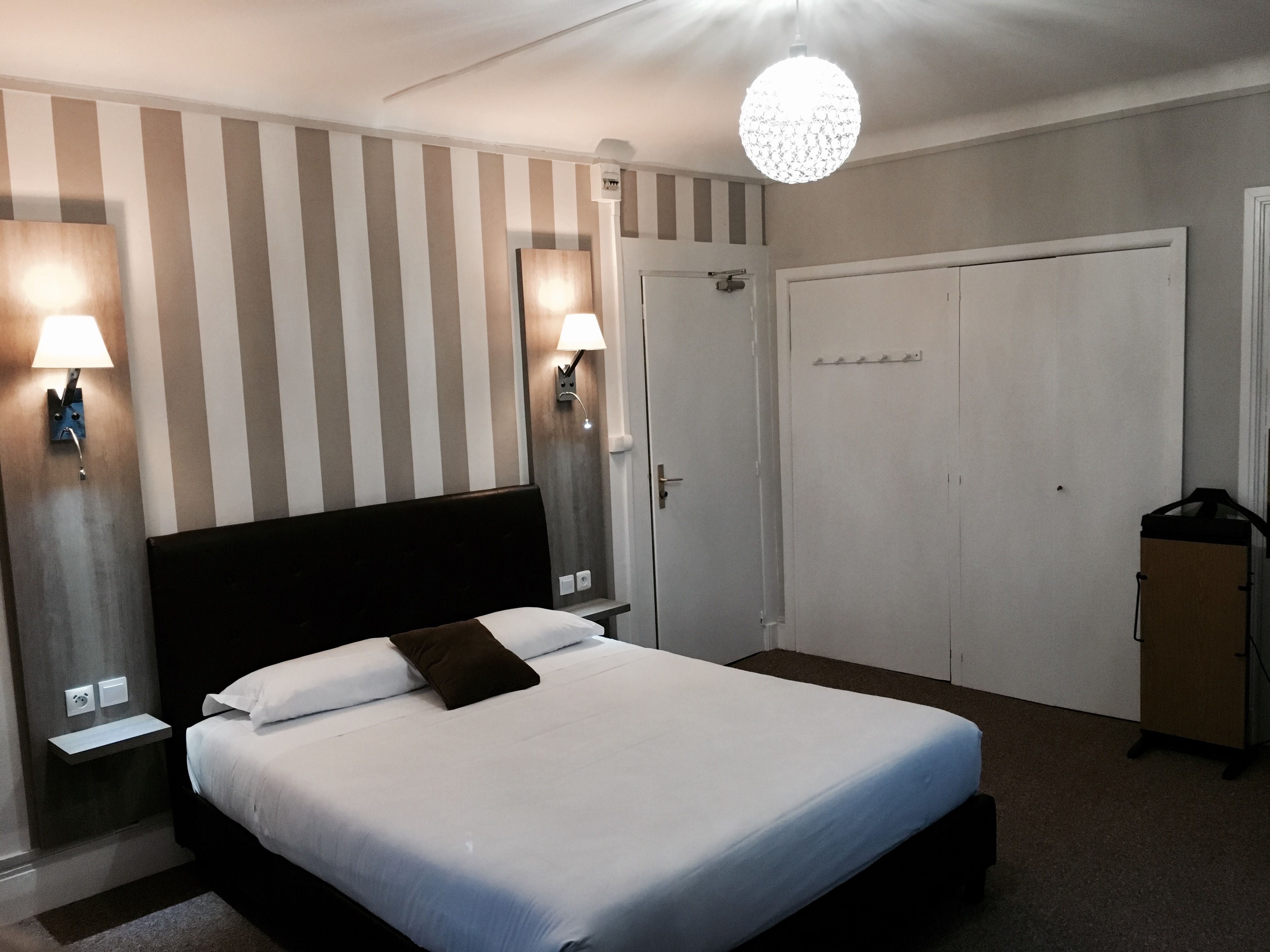 A hotel room with a bed, bedside lamps, a wardrobe, and a hanging light fixture.