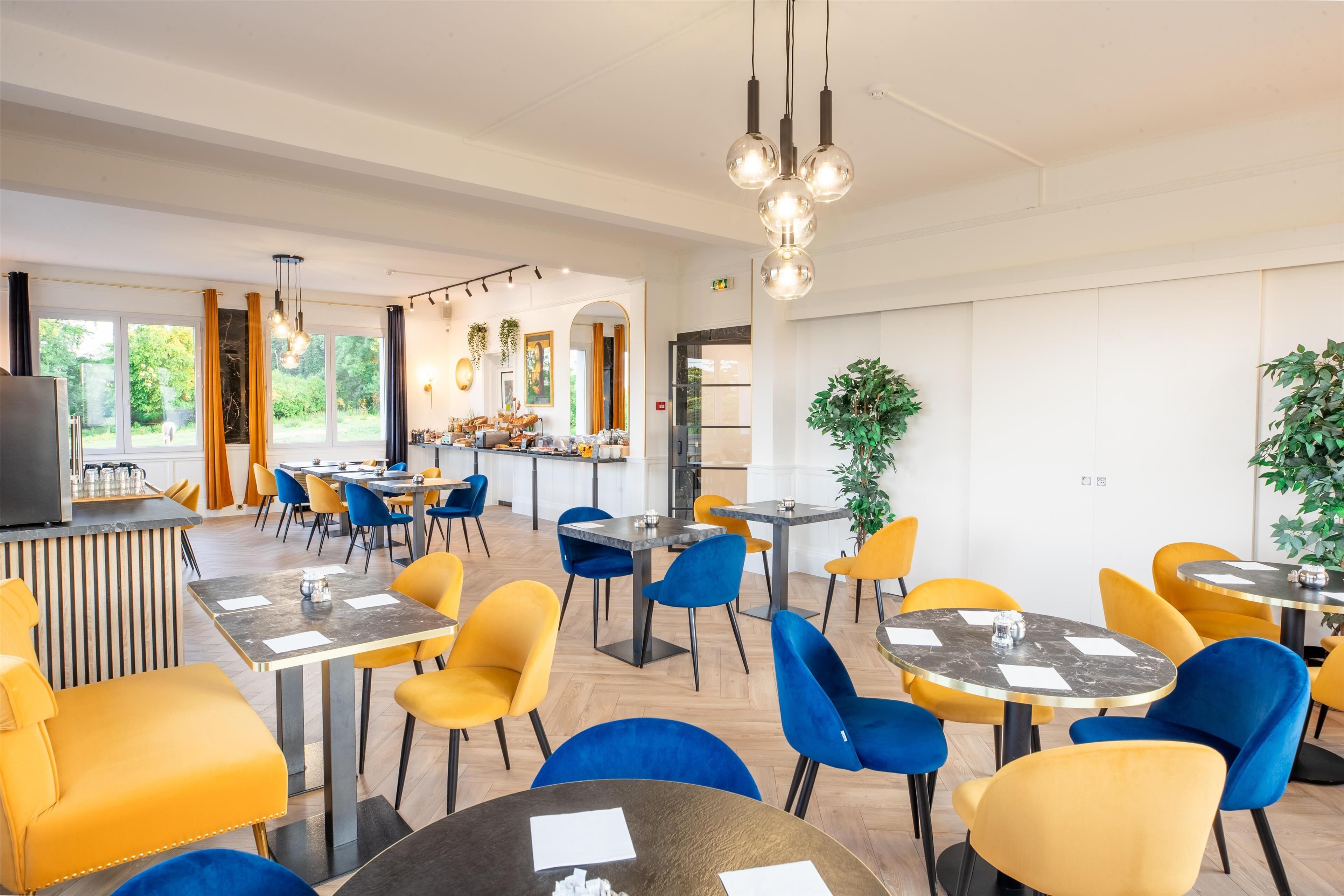 A modern dining area with round tables and vibrant yellow and blue chairs, featuring a kitchen area and large windows.