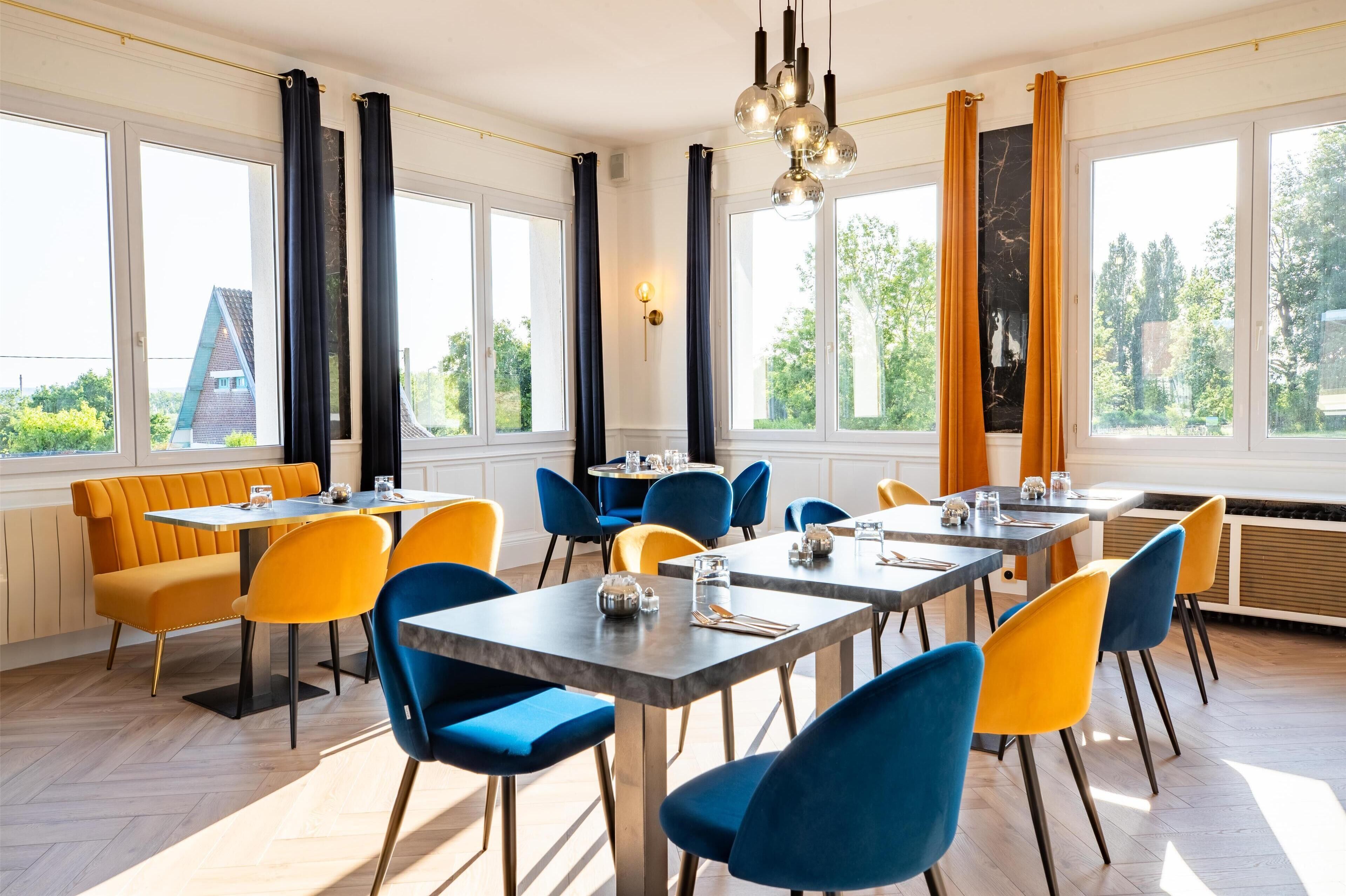 A dining area with tables and chairs, large windows, and a view of greenery outside.