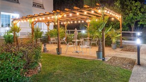 Un patio al aire libre con una pérgola de madera, luces colgantes, plantas en macetas y sillas de metal.