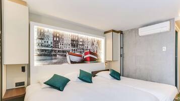 Habitación de hotel con una cama grande, dos almohadas, una mesita de noche, aire acondicionado instalado en la pared y una obra de arte con un paisaje urbano.