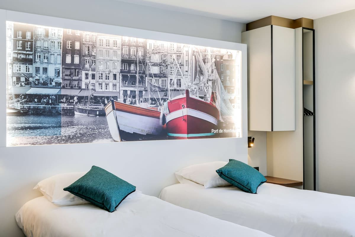 Une chambre d’hôtel avec deux lits, des oreillers de couleur turquoise, un grand tableau encadré représentant un port et une armoire.