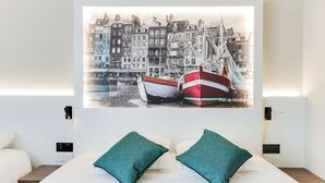 A hotel room with a bed, two teal pillows, a wall-mounted artwork of a harbor scene, and a nightstand with a speaker.