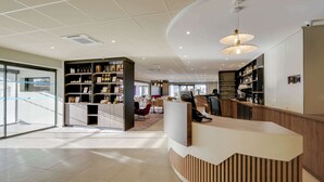 A modern hotel lobby with a reception desk, seating area, and shelves with various items.