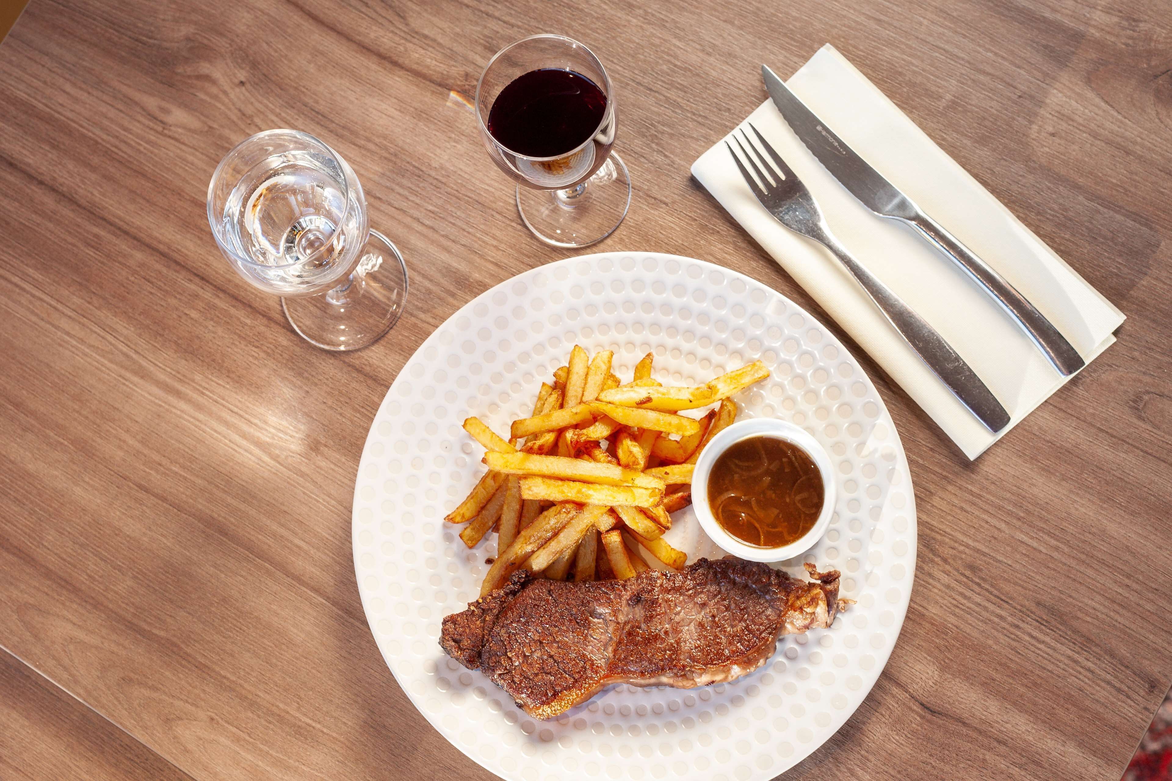 Une assiette avec un steak, des frites et un petit bol de sauce, accompagnée d’un verre d’eau et d’un verre de vin rouge, le tout posé sur une table en bois.