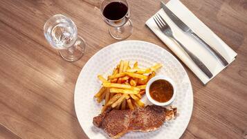 A plate with a steak, fries, and a small bowl of sauce, accompanied by a glass of water and a glass of red wine on a wooden table.