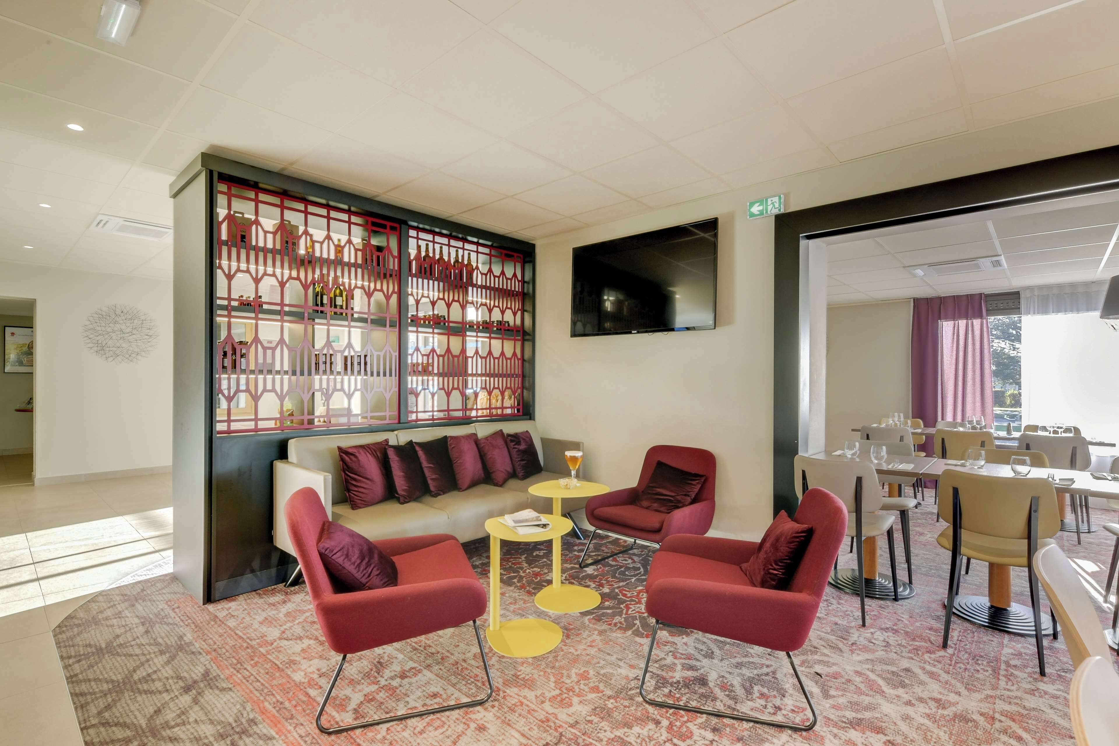 A hotel lobby with a seating area featuring red chairs and a yellow table, a decorative partition with bottles, and a flat-screen TV mounted on the wall.