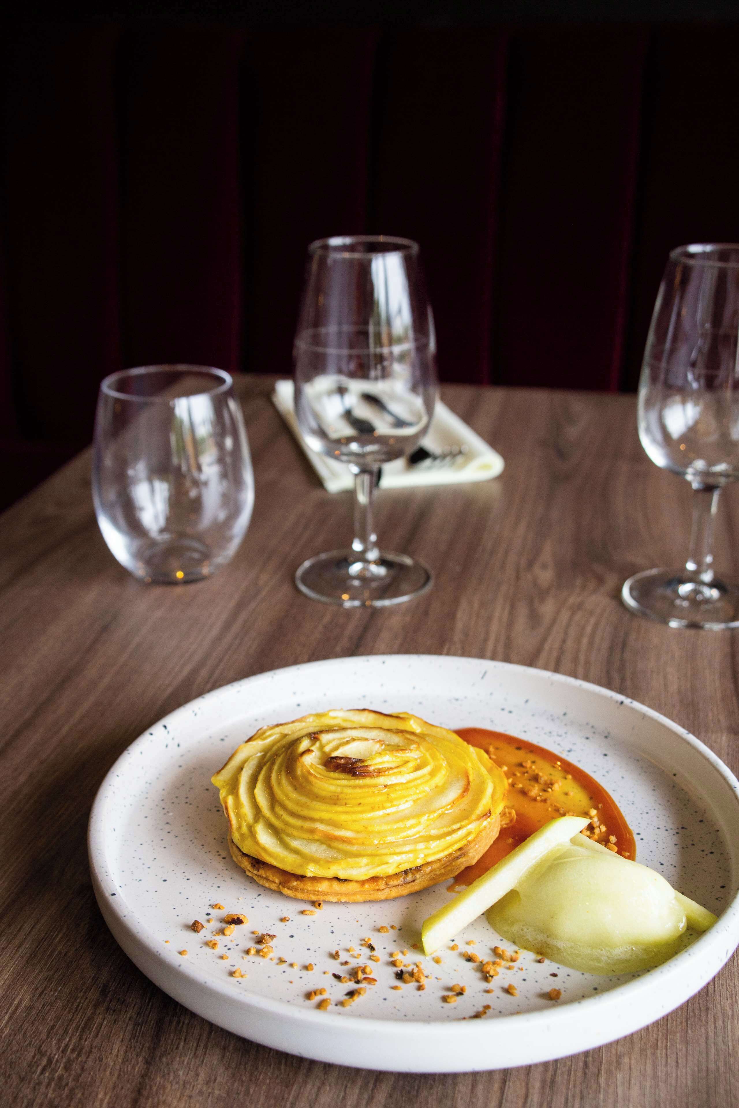 A dessert on a plate with a wooden table and three empty wine glasses in the background.
