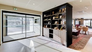 A modern hotel lobby with a glass door entrance, a black bookshelf with wine bottles, and a dining area with tables and chairs.
