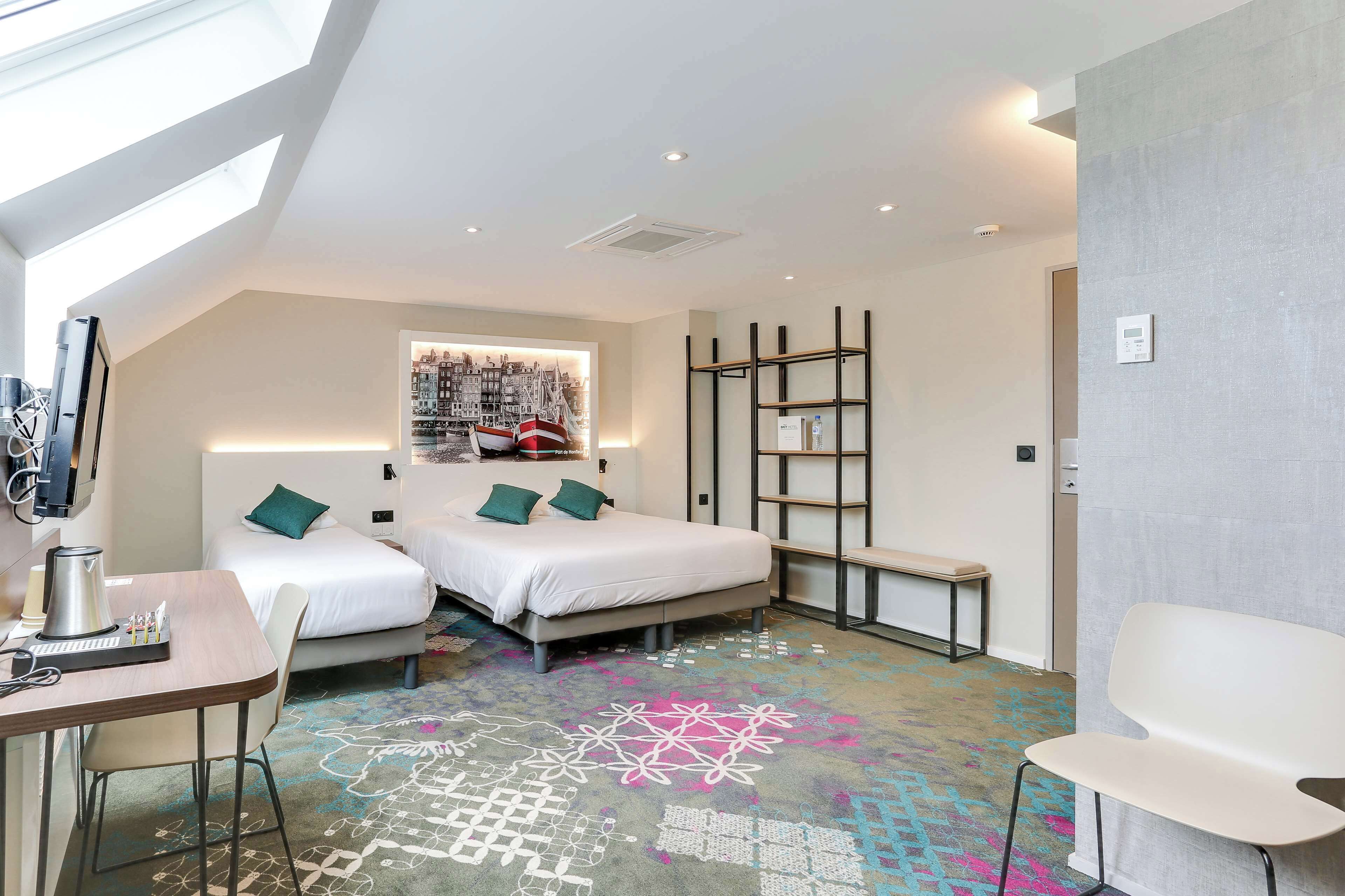 A modern hotel room with a skylight, two beds with green pillows, a desk with a coffee maker, a colorful carpet, and a cityscape wall art.