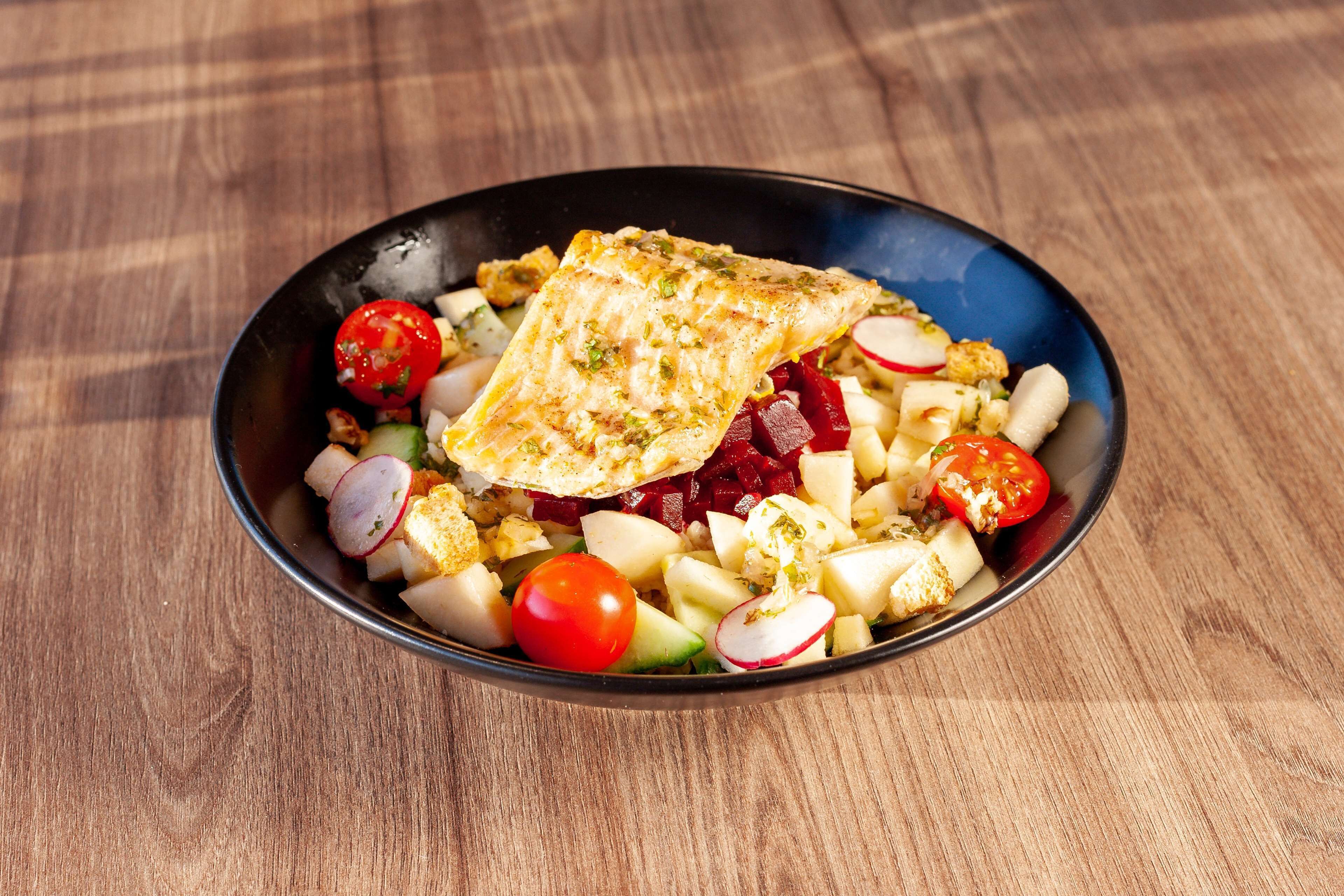 A bowl of salad with various vegetables and a piece of grilled fish on a wooden table.