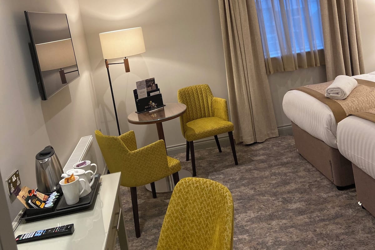 A hotel room with a bed, a desk with a coffee maker, two chairs, a lamp, and a window with curtains.