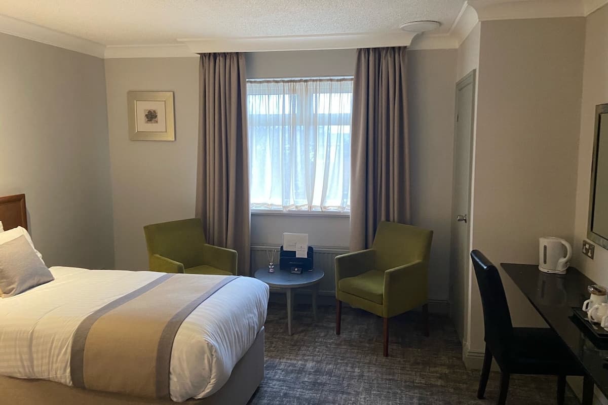 A hotel room with a bed, two chairs, a small table, a window with curtains, a coffee maker, and a framed picture on the wall.