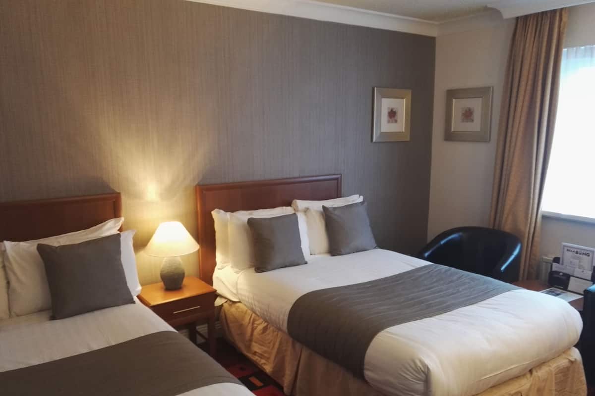 A hotel room with two beds, a nightstand with a lamp, a chair, and a window with curtains.