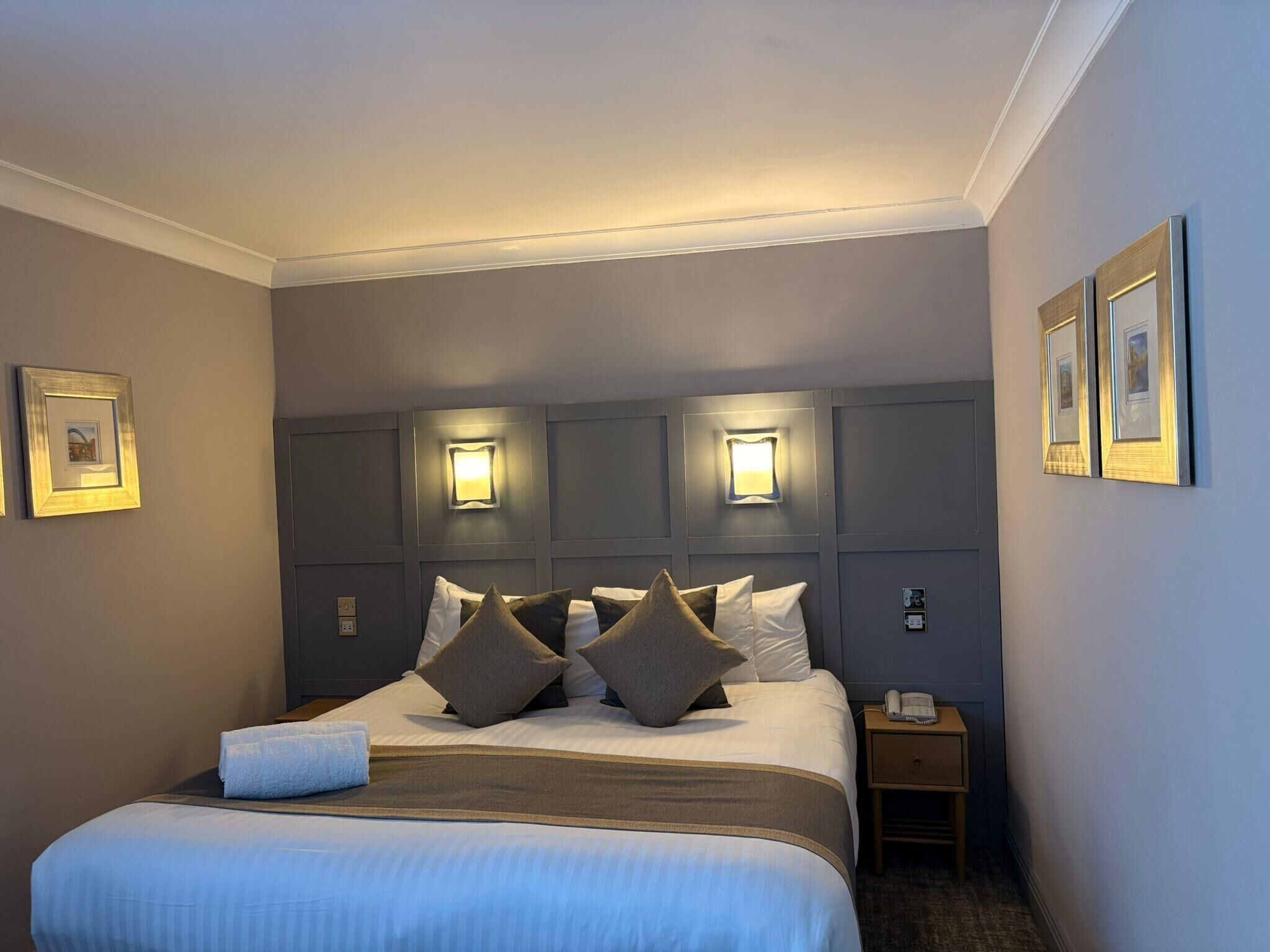 A hotel room with a large bed, a bedside table with a lamp, and framed pictures on the walls.