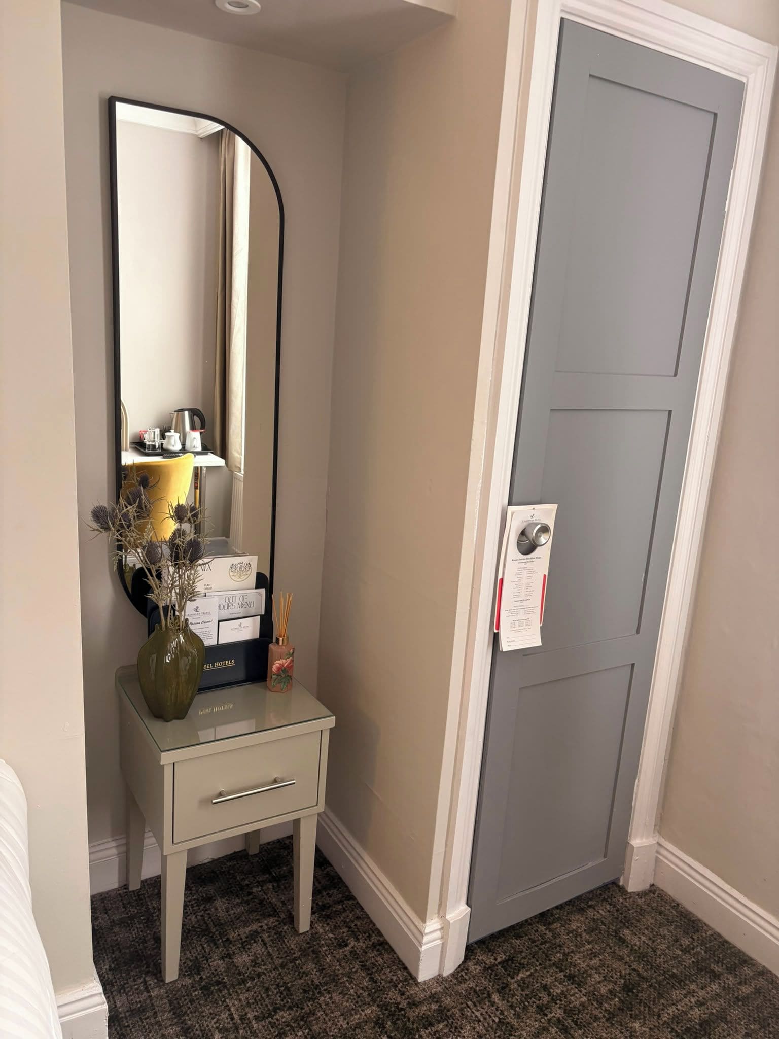Ein Hotelzimmer mit Schminktisch, Spiegel, einer Tür und einem kleinen Tisch mit Vase und Speisekarte.