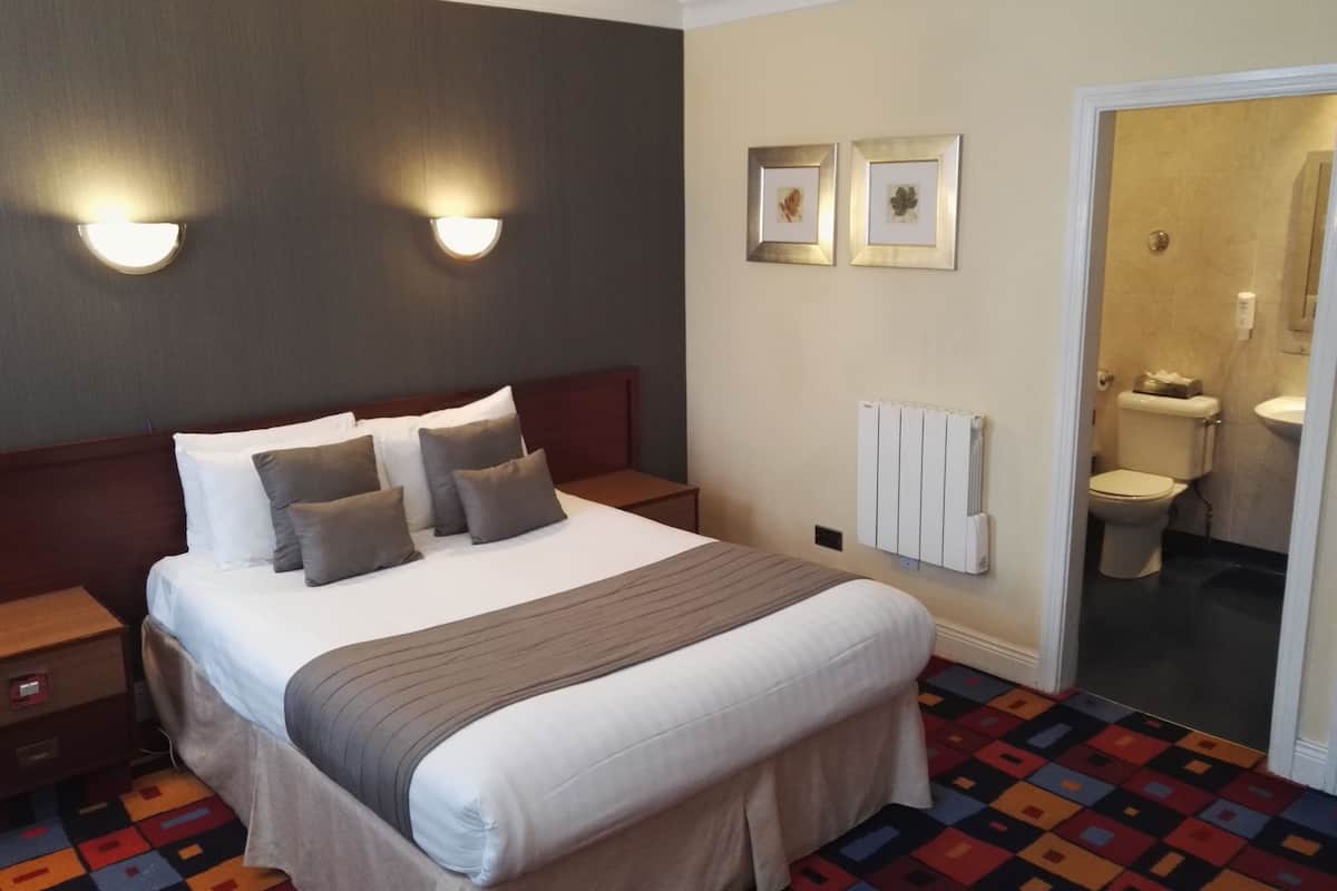 A hotel room with a bed, bedside tables, a radiator, a bathroom with a toilet and sink, and two wall-mounted lights.