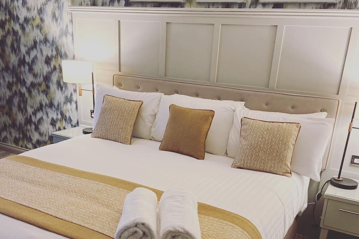 A neatly made bed with a beige and white bedspread, two rolled towels, and a headboard with a panel design.