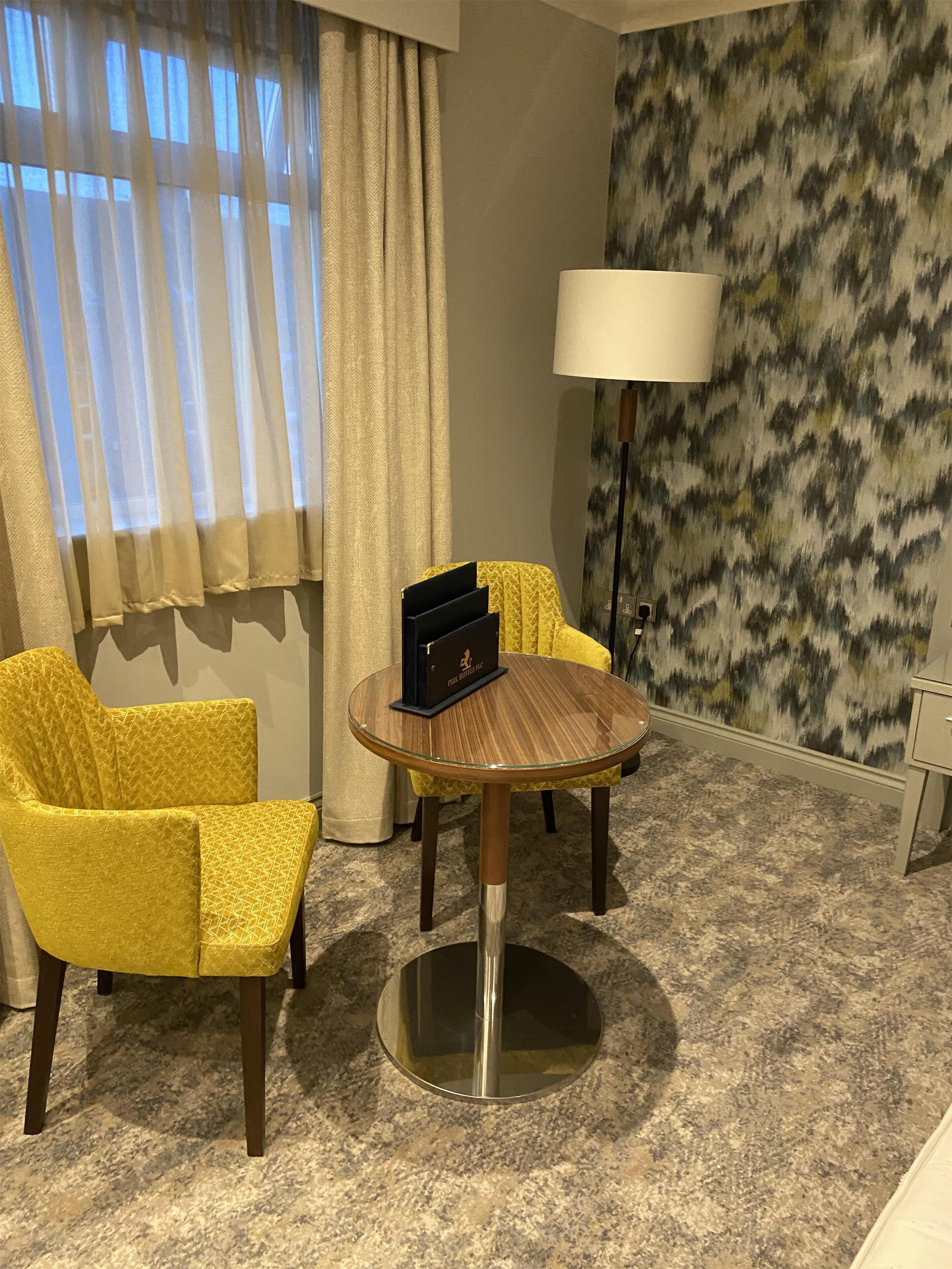 A hotel room with a round table, two yellow armchairs, a lamp, and a patterned wall.