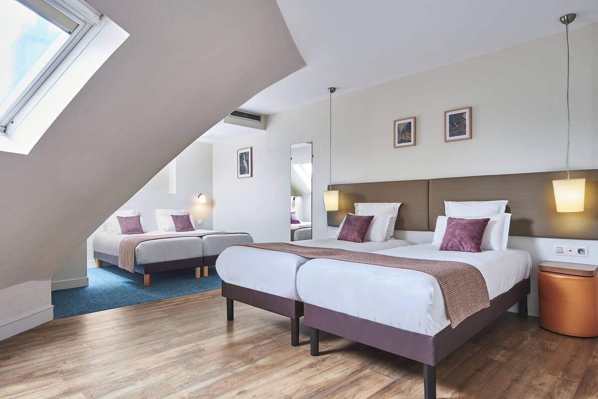 Habitación de hotel moderna con una cama grande, otra más pequeña y un escritorio. Cuenta con una ventana que ofrece luz natural y vistas al cielo.