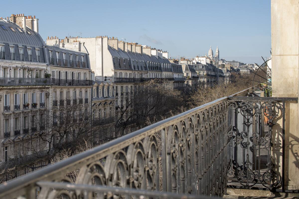 Una hilera de edificios históricos con balcones ornamentados, un río al fondo y un puente con detalles de metalurgia intrincada.