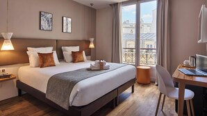 Habitación de hotel con una cama grande, lámparas de noche, un escritorio con una computadora portátil y vistas a edificios a través de la ventana.