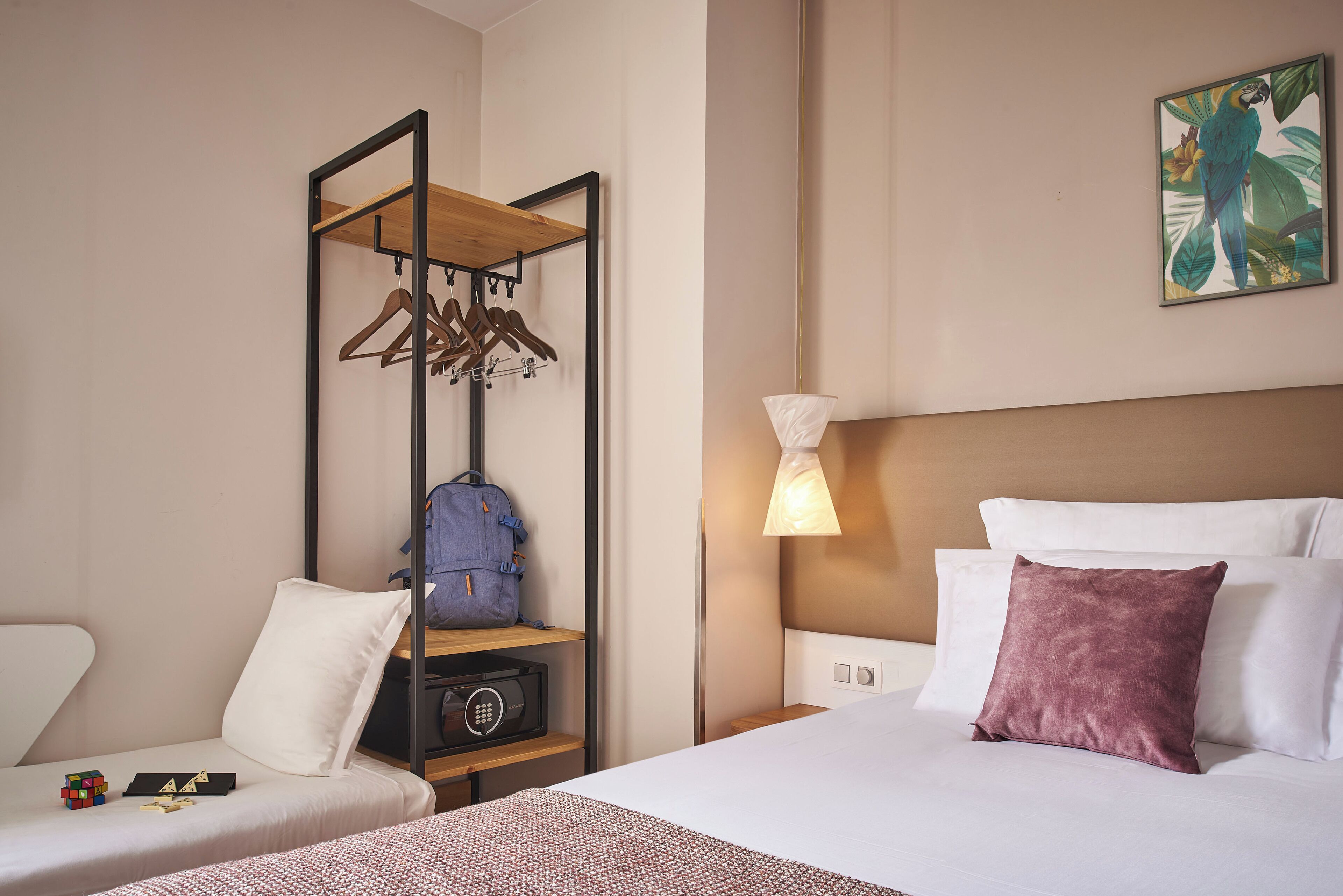 A hotel room with a bed, a side table, a lamp, a hanging coat rack, and a suitcase.