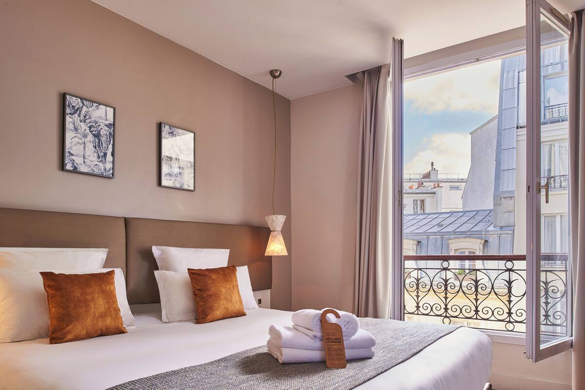 Una habitación de hotel con cama, almohadas, manta, perchero, obra de arte en la pared, ventana con vista a edificios y lámpara colgante.