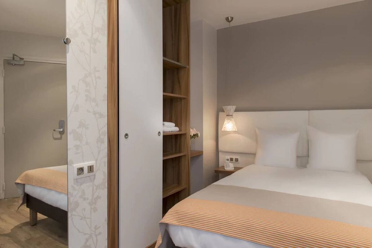 Una habitación de hotel con una cama grande, una mesita de noche con lámpara, un armario y zona de ducha.