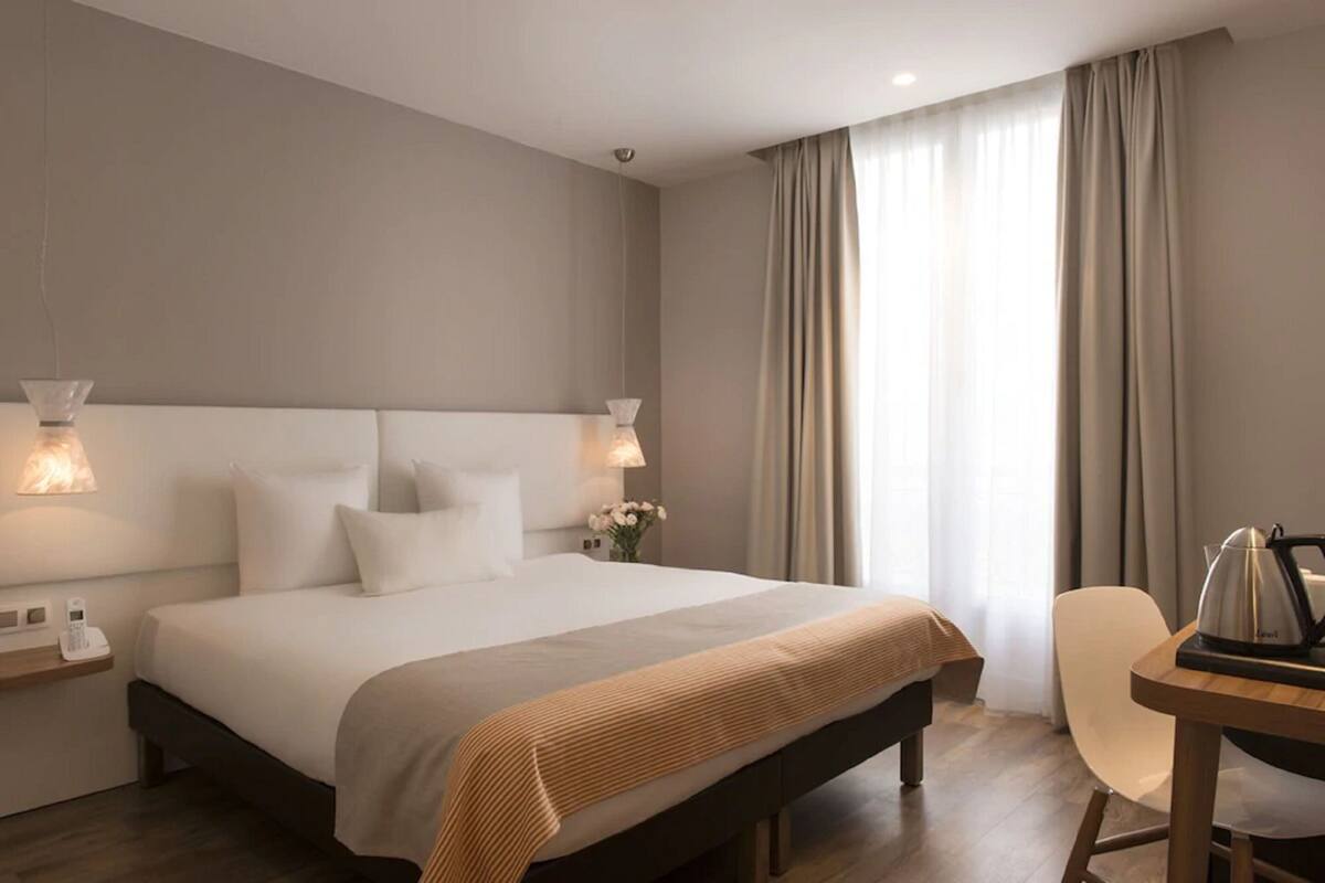 Una habitación de hotel moderna con una cama grande, lámparas de noche, un escritorio con hervidor y vistas al exterior a través de cortinas translúcidas.