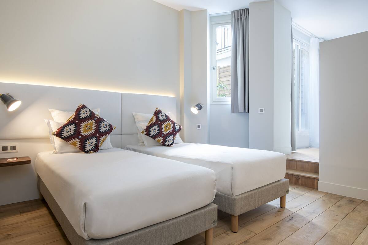 Habitación de hotel moderna con dos camas individuales, una mesita de noche pequeña, un banco integrado, una ventana con cortinas y una lámpara fijada a la pared.