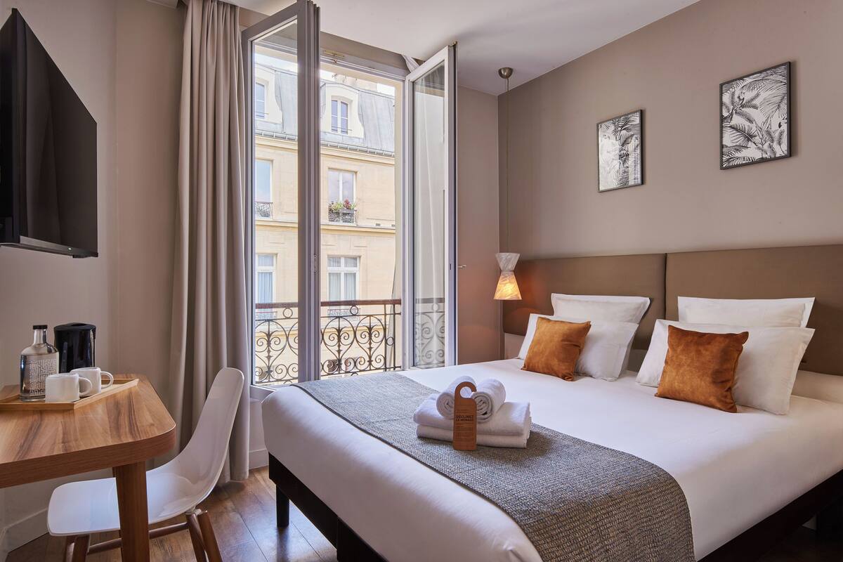 Habitación de hotel con cama, escritorio con silla, cafetera y ventana con cortinas.