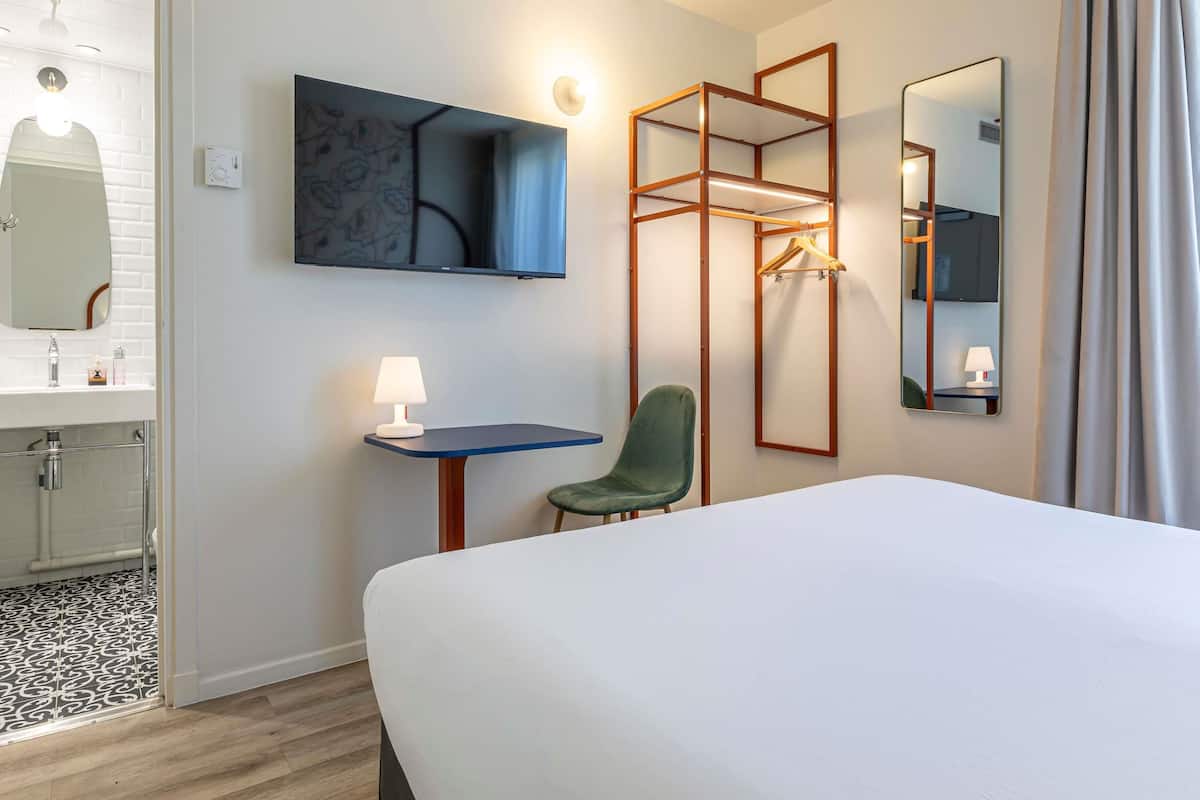 Une chambre d’hôtel comprenant un lit, un bureau avec une lampe, une chaise, une télévision, un miroir et un espace douche.