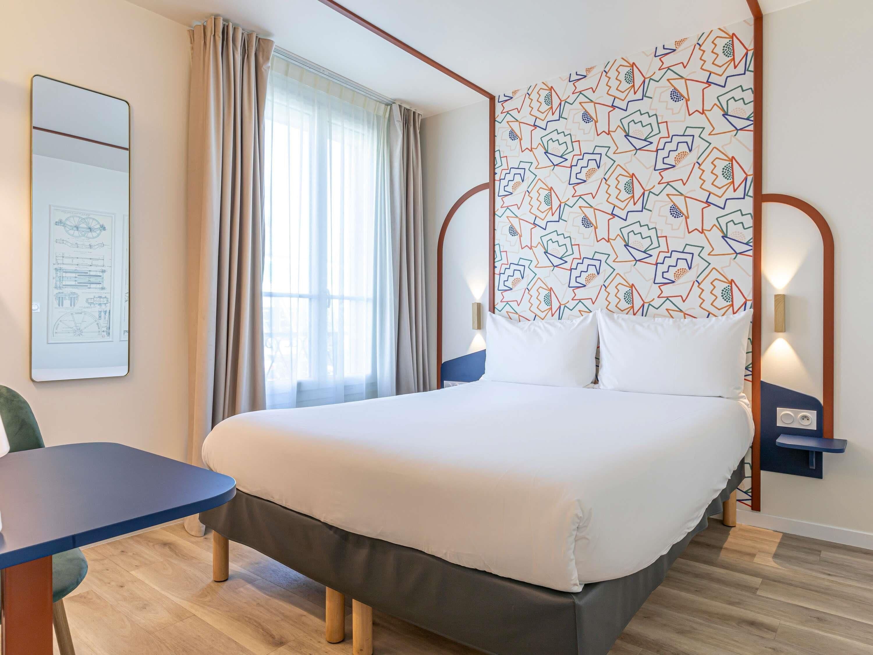 Une chambre d’hôtel comprenant un lit, un bureau, une chaise, une fenêtre avec des rideaux et un mur orné d’un motif géométrique.