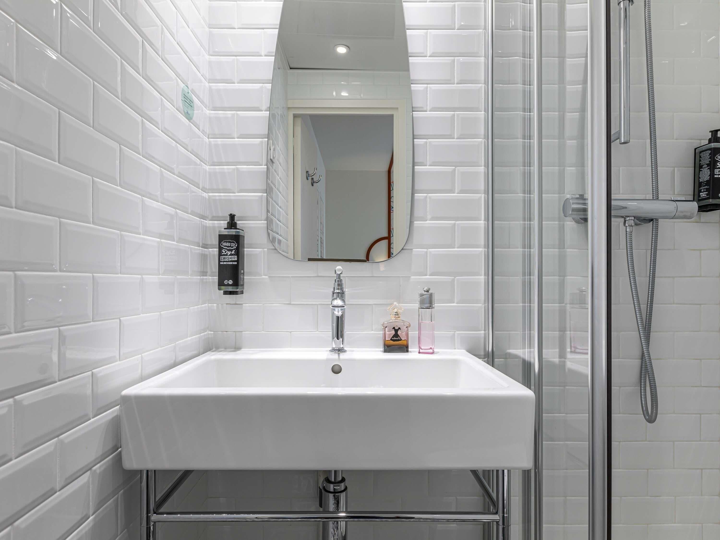 Een badkamer met een witte betegelde muur, een witte wastafel, een spiegel en een douchegedeelte.